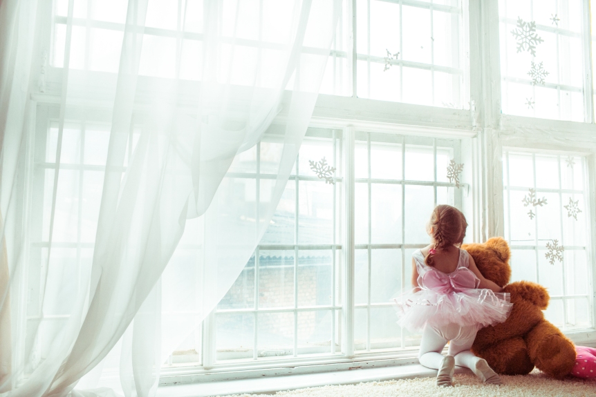 Girl in dancer's suit sits with toy bear before the window